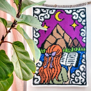 May include: A handmade tapestry featuring a vibrant scene of a person with orange hair reading a book under a purple sky with mountains, a crescent moon, and stars. The tapestry is framed with white clouds and a black border.