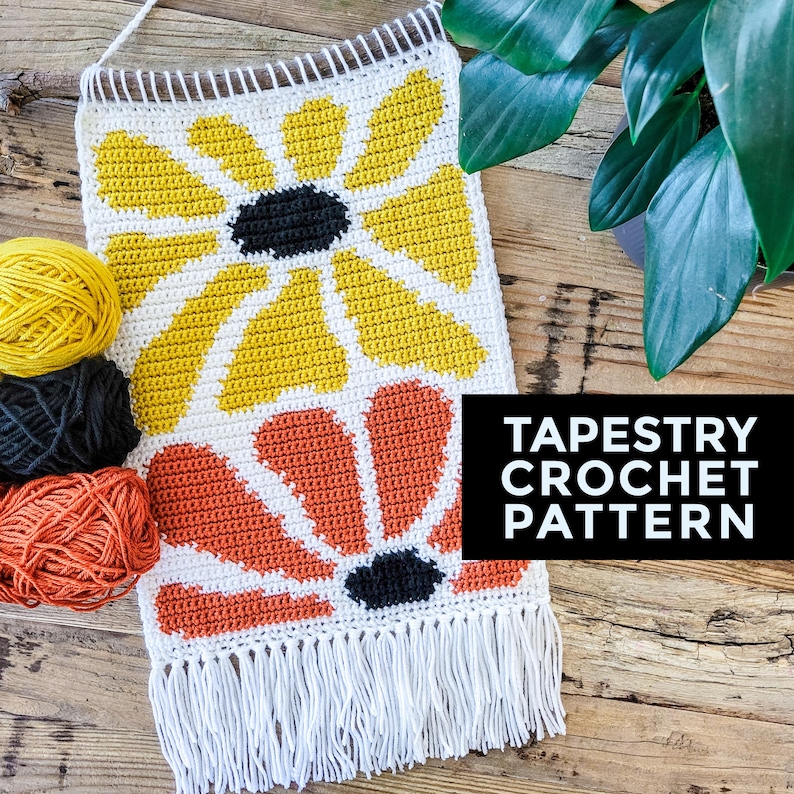 Pu&ograve; includere: Uno schema di arazzo all'uncinetto con due grandi fiori, uno giallo e uno arancione, con centri neri. L'arazzo ha un bordo di frange bianche e il testo "TAPESTRY CROCHET PATTERN" &egrave; visualizzato sotto l'immagine.