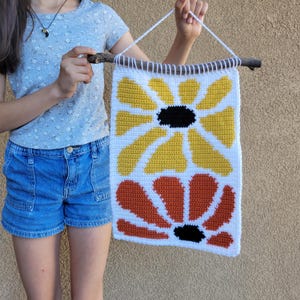 Pu&ograve; includere: Un arazzo da parete all'uncinetto con due grandi fiori. Il fiore superiore &egrave; giallo con un centro nero e il fiore inferiore &egrave; arancione con un centro nero. I fiori sono all'uncinetto su uno sfondo bianco.