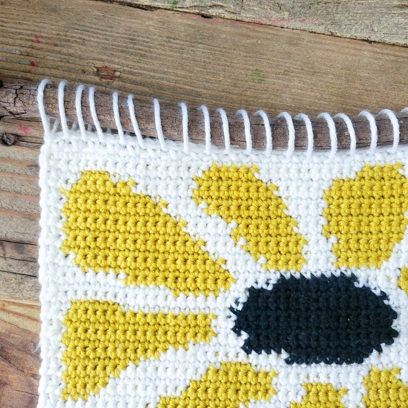Pu&ograve; includere: Un arazzo da parete all'uncinetto con un girasole giallo con un centro nero. Il girasole &egrave; lavorato all'uncinetto con un semplice motivo a punti ed &egrave; incorniciato da un bordo bianco.