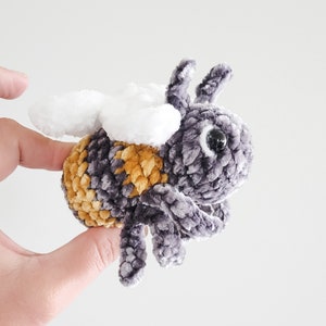 May include: A plush bee toy with a gray fuzzy body, yellow and brown stripes, and white wings. The bee has large black eyes.