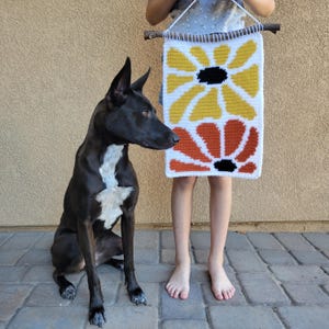 Pu&ograve; includere: Un cane bianco e nero siede di fronte a un arazzo da parete all'uncinetto con due grandi fiori colorati. I fiori sono gialli e arancioni con centri neri.