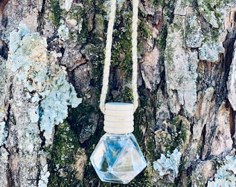 May include: A clear glass bottle with a geometric shape hangs from a white string with a wooden bead at the top. The bottle is filled with a clear liquid and is hanging against a tree trunk.