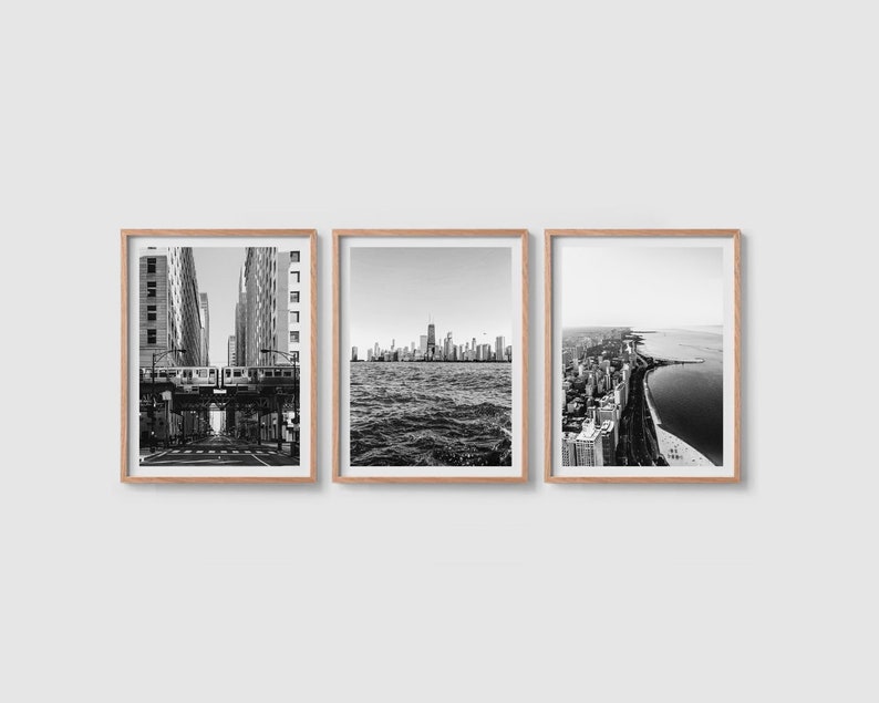 May include: Set of three framed black and white photographs. The images depict urban cityscapes, including a train on an elevated track, a skyline over water, and a coastal view with buildings and a beach. The frames are a light wood color.
