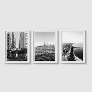 May include: Set of three framed black and white photographs. One shows a city street with a train, another a skyline over water, and the third an aerial view of a city and shoreline. The frames are white.