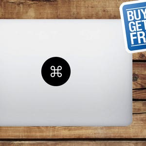 May include: A silver laptop with a black circle sticker featuring a white command symbol. A blue sticker in the upper right corner reads "BUY 1 GET FREE". The laptop is on a wooden surface.