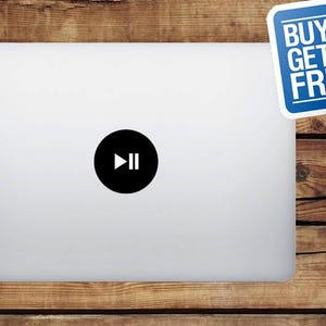 May include: A silver laptop with a black circle containing a white play and pause symbol. A blue sticker in the upper right corner reads "BUY 1 GET FREE". The laptop is on a wooden surface.