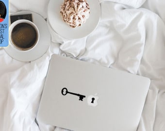 May include: A silver laptop with a black key and keyhole decal. The key is next to the Apple logo. A cup of coffee, a plate with a pastry, and a sign that says "LONDON'S FINEST DECAL SHOP" are also in the image.