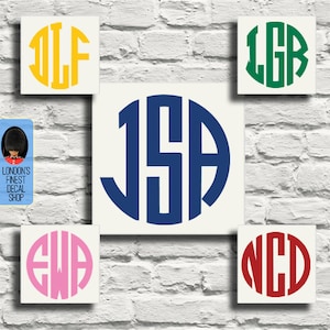 Op de afbeelding: Een verzameling monogramstickers in verschillende kleuren, waaronder geel, groen, roze en rood, weergegeven op vierkante, gebroken witte achtergronden. De monogrammen bevatten drie hoofdletters in een cirkelvormig ontwerp, tegen een witte bakstenen muur.