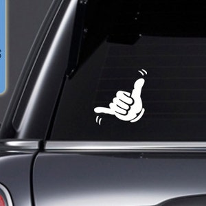 May include: White vinyl decal of a cartoon hand making the shaka sign, a popular Hawaiian gesture of peace, aloha, and good vibes. The decal is on the back window of a black car.
