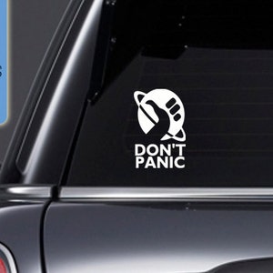 Op de afbeelding: Witte vinyl sticker met een gestileerde hand die een planeet met een ring eromheen vasthoudt. De tekst "DON'T PANIC" staat onder de afbeelding.