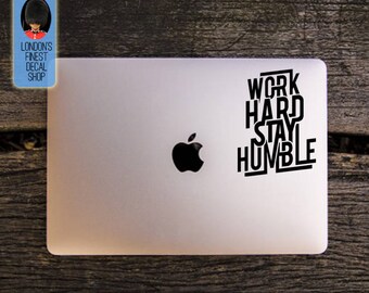 May include: A silver laptop with a black Apple logo and a black vinyl decal that reads "WORK HARD STAY HUMBLE." The decal is in a bold, sans-serif font. A small blue shop logo is in the upper left corner.