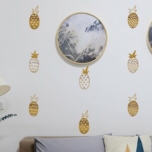 Puede incluir: Una pared blanca decorada con calcomanías doradas de piñas. Dos obras de arte redondas enmarcadas con diseños abstractos cuelgan de la pared. También se ve una lámpara de pie con una pantalla blanca y un sofá con cojines grises.