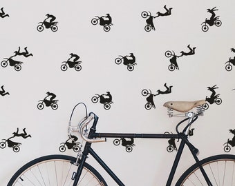May include: A white wall with black silhouette decals of a person performing a stunt on a dirt bike. The decals are repeated in a pattern across the wall. A black bicycle is leaning against the wall.