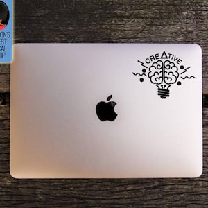 May include: A black vinyl decal featuring a brain inside a lightbulb with the word "CREATIVE" above it, applied to a silver laptop. The laptop has a black Apple logo. The decal is a design from London's Finest Decal Shop.