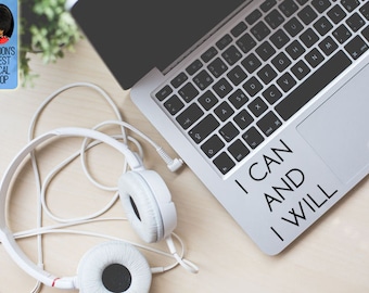 May include: A silver laptop with a black keyboard and the words "I CAN AND I WILL" in black. White headphones and a blue sticker with "LONDON'S FINEST DECAL SHOP" are on a light wooden surface.
