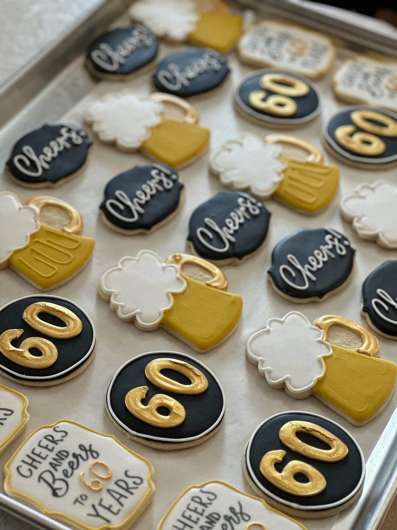 May include: A collection of decorated sugar cookies with various designs, including beer mugs, the number 60, and the phrase "Cheers and Beers to 60 Years". The cookies are arranged on a baking sheet.