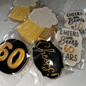 May include: Four decorated sugar cookies. One cookie is black with gold lettering that says "Cheers!" Another cookie is white with gold lettering that says "Cheers and Beers to 60 Years". The other two cookies are yellow with white frosting and a gold outline of a beer mug.