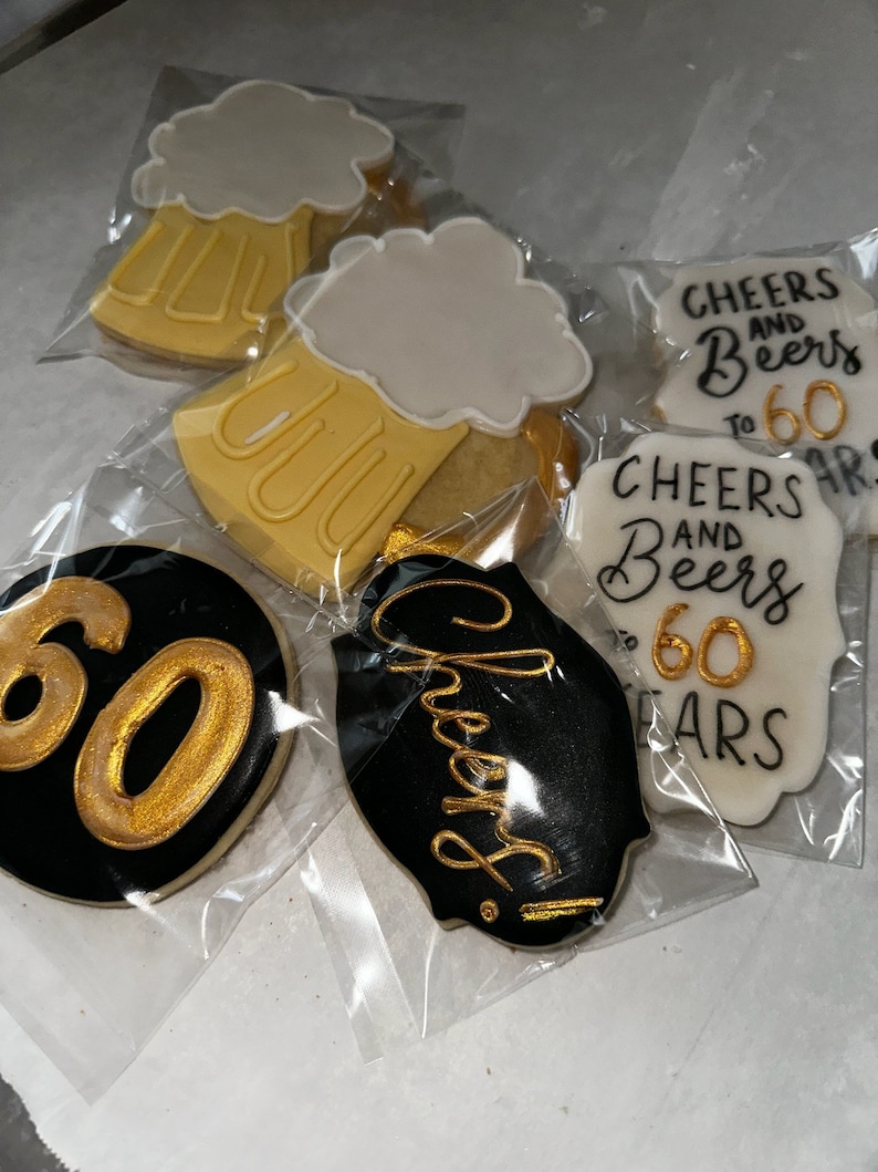May include: Four decorated sugar cookies. One cookie is black with gold lettering that says "Cheers!" Another cookie is white with gold lettering that says "Cheers and Beers to 60 Years". The other two cookies are yellow with white frosting and a gold outline of a beer mug.