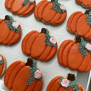 May include: A collection of orange pumpkin-shaped cookies decorated with green leaves and pink flowers. The cookies are arranged on a white surface.