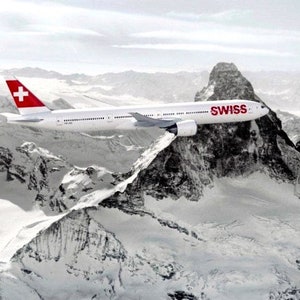 K&ouml;nnte beinhalten: Ein wei&szlig;es Flugzeug mit einer roten Schweizer Flagge am Heck fliegt &uuml;ber eine schneebedeckte Bergkette. Das Flugzeug tr&auml;gt den Schriftzug "SWISS" an der Seite.