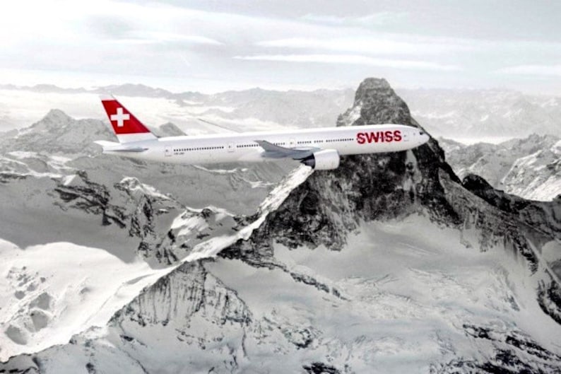 K&ouml;nnte beinhalten: Ein wei&szlig;es Flugzeug mit einer roten Schweizer Flagge am Heck fliegt &uuml;ber eine schneebedeckte Bergkette. Das Flugzeug tr&auml;gt den Schriftzug "SWISS" an der Seite.