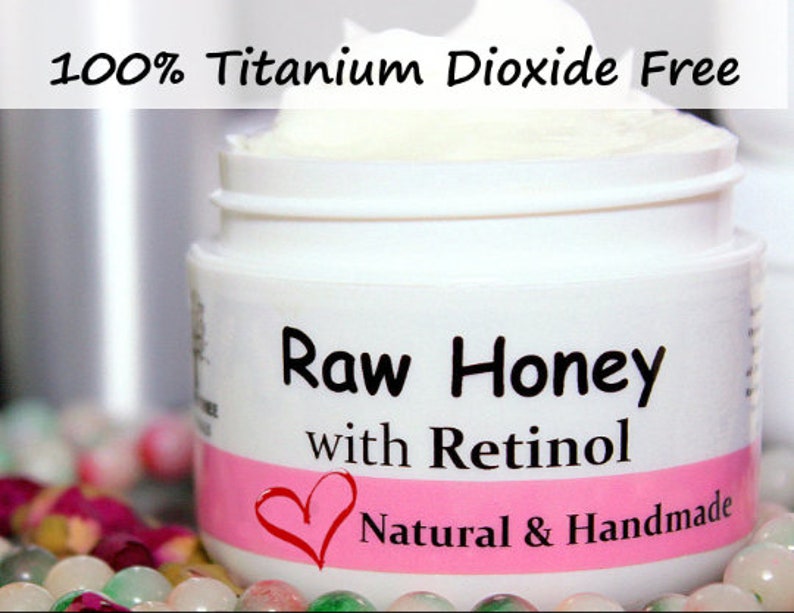 May include: A white jar with a pink label that reads "Raw Honey with Retinol" and "Natural & Handmade". The jar is filled with a white cream and has a white lid. The jar is sitting on a bed of colorful stones.