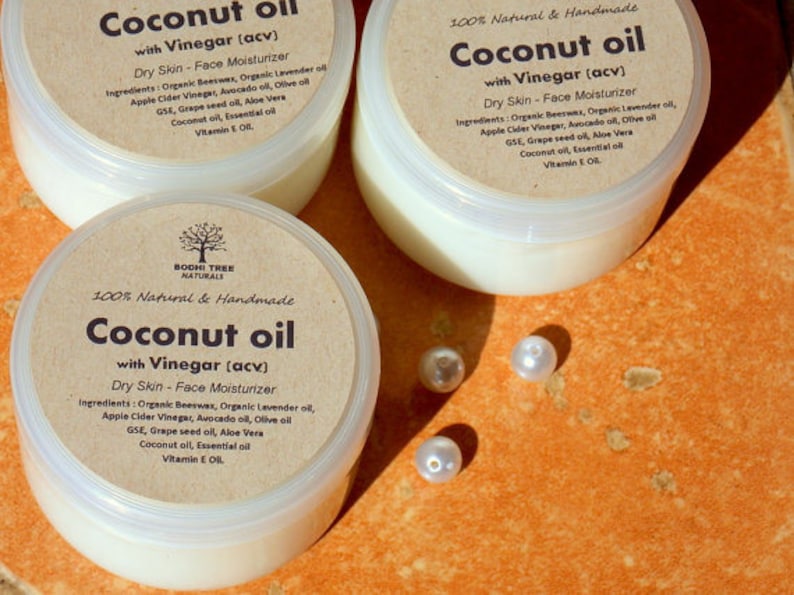 May include: Three glass jars of coconut oil with vinegar, labeled "Coconut oil with Vinegar (acv)" and "100% Natural & Handmade". The jars are on a textured orange surface with white pearl beads scattered around them.