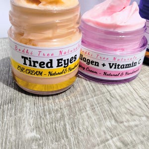 Op de afbeelding: Twee potjes oogcr&egrave;me worden getoond. E&eacute;n potje bevat een perzikcr&egrave;me met het label "Tired Eyes Eye Cream - Natural & Handmade". Het andere potje bevat roze cr&egrave;me met het label "Collagen + Vitamin C - Natural & Handmade."