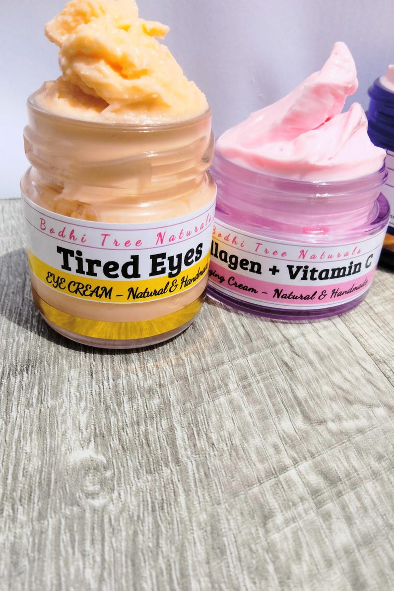 Op de afbeelding: Twee potjes oogcr&egrave;me worden getoond. E&eacute;n potje bevat een perzikcr&egrave;me met het label "Tired Eyes Eye Cream - Natural & Handmade". Het andere potje bevat roze cr&egrave;me met het label "Collagen + Vitamin C - Natural & Handmade."