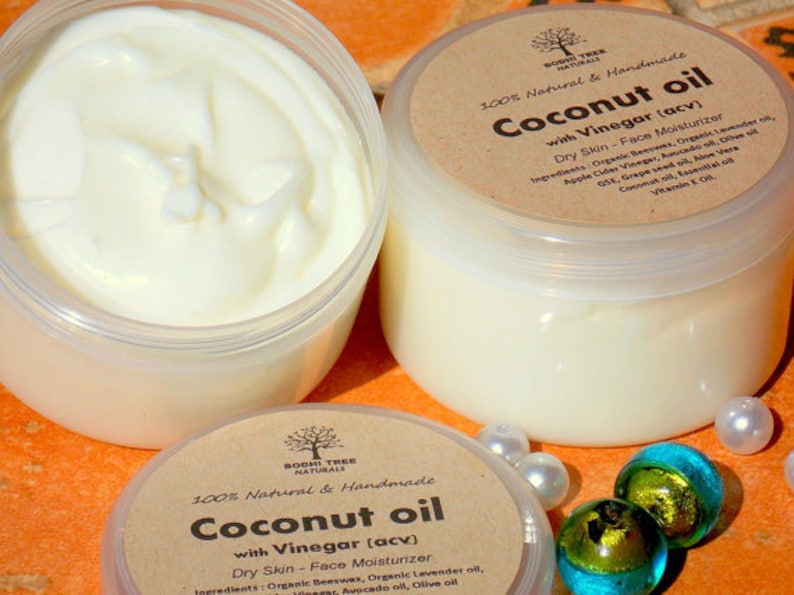 May include: Two glass jars of coconut oil with vinegar face moisturizer. The jars are labeled "100% Natural & Handmade Coconut oil with Vinegar (acv) Dry Skin - Face Moisturizer" and list the ingredients. The jars are sitting on a brown surface with white and green decorative stones.