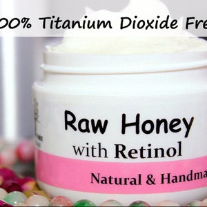 May include: A white jar with a pink label that reads "Raw Honey with Retinol" and "Natural & Handmade". The jar is filled with a white cream and the top of the jar has the text "100% Titanium Dioxide Free".