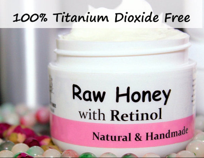 May include: A white jar with a pink label that reads "Raw Honey with Retinol" and "Natural & Handmade". The jar is filled with a white cream and the top of the jar has the text "100% Titanium Dioxide Free".