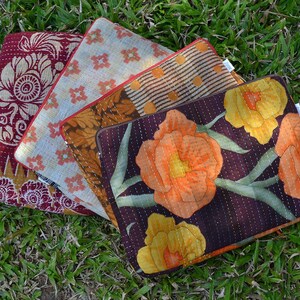 May include: Four colorful quilted laptop sleeves with different patterns. The sleeves are made of fabric with floral designs, geometric patterns, and a brown and white striped pattern. The sleeves are arranged in a stack, with the floral sleeve on top.