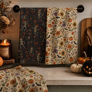 May include: Two rectangular kitchen towels hanging on a rod and one folded on a counter. One towel features a dark background with dancing skeletons and flowers. The other two towels feature a cream background with pumpkins, flowers, and bats.