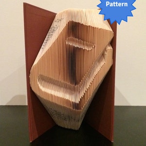 Könnte beinhalten: Ein Buch, das zu einer dreidimensionalen Skulptur des Buchstabens "I" gefaltet ist. Das Buch ist in braunes Leder gebunden und die Seiten sind nach einem Muster gefaltet. Die Skulptur steht auf einer schwarzen Oberfläche. Das Wort "Pattern" steht auf einem blauen Stern.