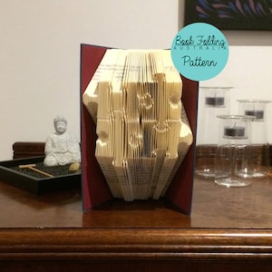 Puede incluir: Un libro doblado en forma de casa, con las palabras "Book Folding Australia Pattern" en un círculo azul. El libro está sobre una mesa de madera con una pequeña estatua de Buda y una bandeja con un rastrillo.