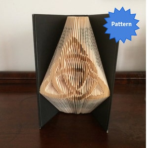 May include: A book folded into a three-dimensional sculpture of a triangle with a pattern inside. The book is standing upright on a wooden surface. The sculpture is made from the pages of a book, and the pages are folded to create the shape. The sculpture is a light brown color. The word "Pattern" is written in blue on a blue starburst.