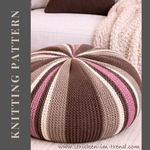 May include: A round, striped knitted pouf in shades of brown, pink, and cream. The pouf is on a light-colored sofa with a matching knitted throw blanket and a brown pillow. The text "KNITTING PATTERN" is on the left side of the image.