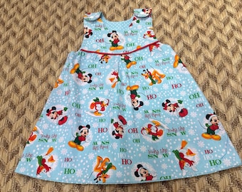 May include: A light blue sleeveless dress featuring a repeating pattern of Mickey Mouse, Pluto, and Donald Duck in holiday attire, interspersed with white snowflakes and the text "OH HO HO" and "looks like SNOW" in red and green.