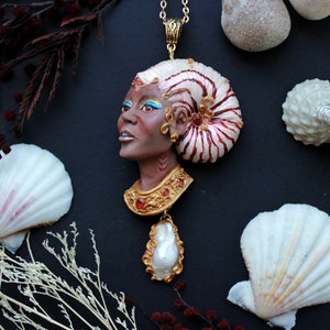 May include: A gold necklace with a pendant featuring a sculpted face with a nautilus shell headdress. The pendant is adorned with red and gold accents. The necklace is displayed on a black background with seashells and dried flowers.