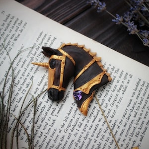 Peut inclure: Une sculpture de tête de licorne noire et dorée avec une pierre précieuse violette. La licorne porte une bride dorée et a une corne. La sculpture est sur un livre avec du texte.