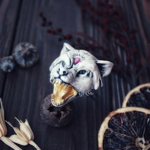 May include: A white ceramic wolf head ring with a gold-painted mouth and a pink gemstone on the forehead. The ring is on a dark brown wooden surface.