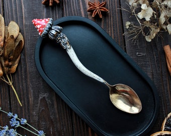 May include: A white and red mushroom-shaped spoon with a gold handle on a black oval tray. The spoon is on a wooden surface with dried flowers and cinnamon sticks.