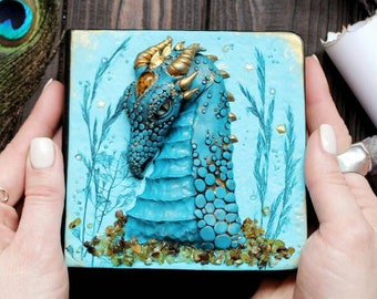 May include: A small, blue and gold dragon sculpture is displayed on a blue background with gold accents. The sculpture is surrounded by small, white flowers and gold glitter. The sculpture is held in the hands of a person.