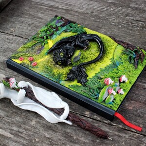 May include: A black and green notebook with a 3D sculpted dragon on the cover. The dragon is lying down with its wings spread out. The notebook has a red ribbon bookmark. A white and brown pen with a sculpted dragon head is lying next to the notebook.