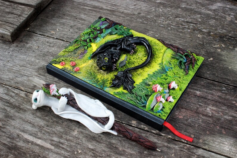 May include: A black and green notebook with a 3D sculpted dragon on the cover. The dragon is lying down with its wings spread out. The notebook has a red ribbon bookmark. A white and brown pen with a sculpted dragon head is lying next to the notebook.
