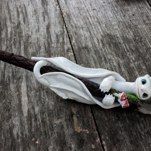May include: White dragon figurine with blue eyes holding a brown stick with green and red accents. The dragon is curled around the stick, which is on a wooden surface.