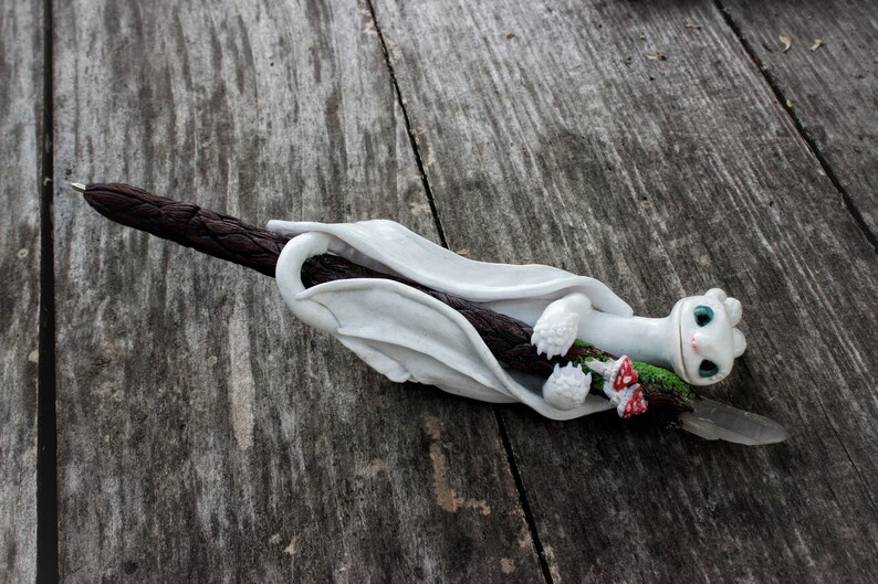 May include: White dragon figurine with blue eyes holding a brown stick with green and red accents. The dragon is curled around the stick, which is on a wooden surface.