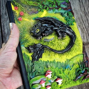 May include: A black notebook with a 3D sculpted black dragon on the cover. The dragon is lying on a green grassy field with red and white mushrooms. The notebook has a black spine and a red ribbon bookmark.